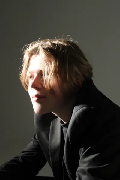 Head-and-shoulders portrait of J.J. Barton with hair falling over one eye, in a dark jacket, side-lit.