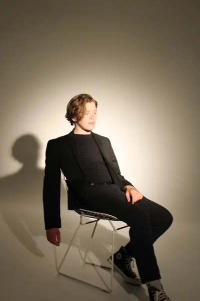 J.J. Barton leaning back on a wire chair with his eyes closed, in an all-black suit, a long shadow behind.
