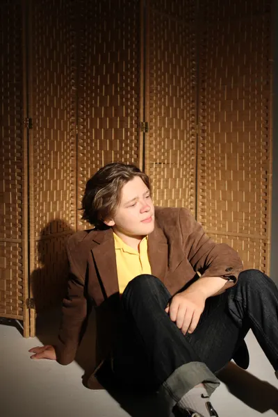 J.J. Barton seated on the floor with one knee drawn up, in a brown jacket and yellow polo, a woven screen behind him.