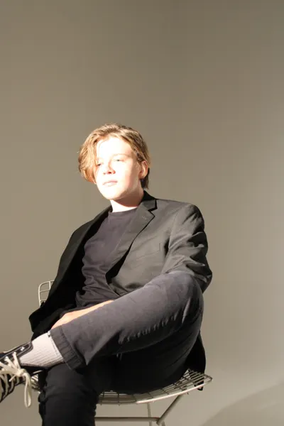 J.J. Barton seated with legs crossed, looking to camera, in a dark blazer and jeans.