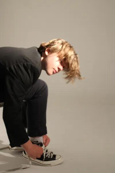 J.J. Barton bending down to tie the lace of a black canvas high-top, hair falling across his face.