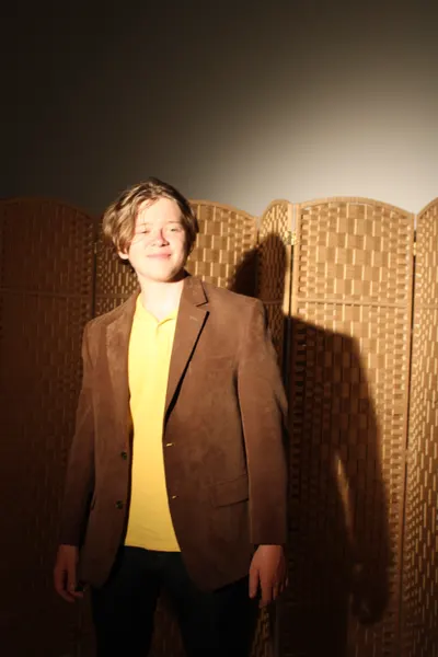 J.J. Barton smiling at the camera in a brown jacket and yellow polo, standing in front of a woven screen.