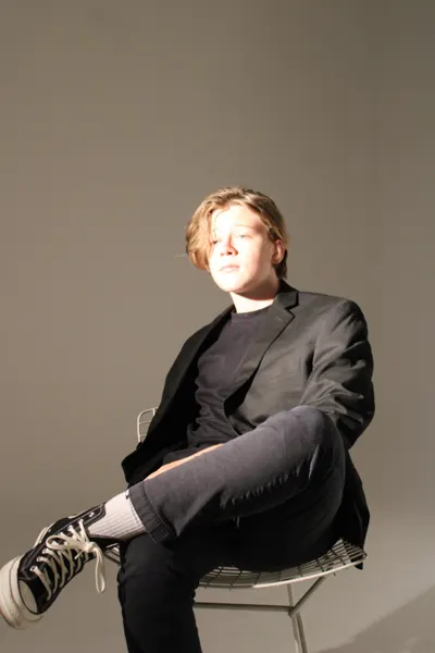 J.J. Barton sitting on a wire chair with his legs crossed, one high-top sneaker toward the camera.