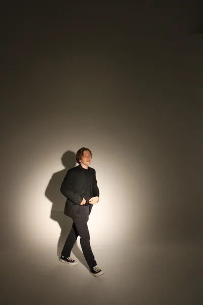 Full-length shot of J.J. Barton in a dark suit mid-stride, casting a long shadow across a spotlit studio wall.