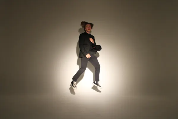 J.J. Barton caught mid-jump in a dark suit and canvas high-tops, arms drawn in, against a spotlit backdrop.