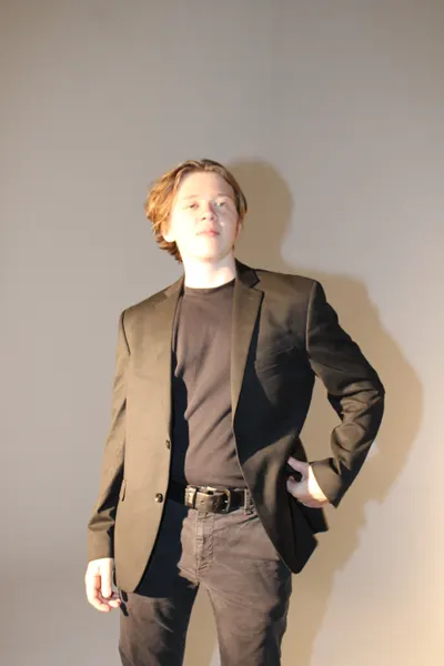 J.J. Barton standing with a hand on his hip in an olive-brown blazer, chin raised toward the camera.