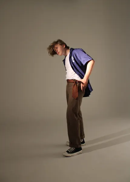 Full-length studio shot of J.J. Barton standing with hands behind his back, head tipped to one side, in a blue shirt and brown trousers.