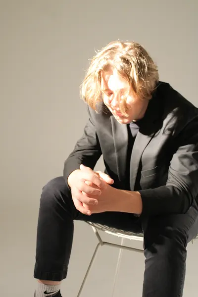 J.J. Barton sitting on a wire chair with his hands clasped, head bowed, in a dark suit.
