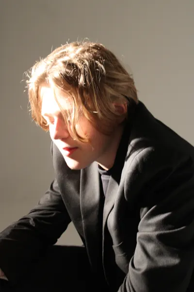 J.J. Barton looking down with his hair falling forward, in a dark jacket, under a hard side light.