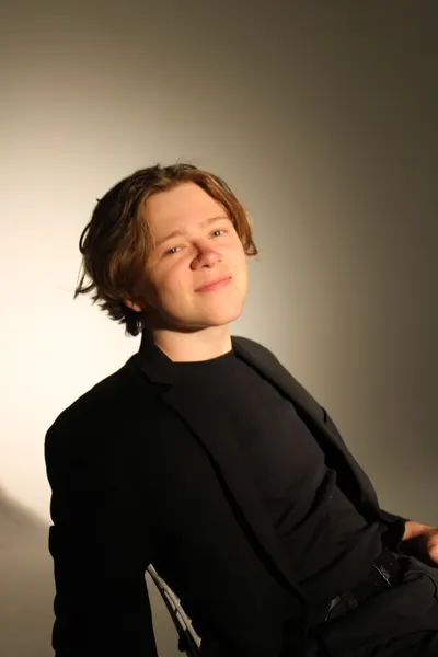 J.J. Barton seated, head tilted to one side, smiling faintly at the camera in a black suit.
