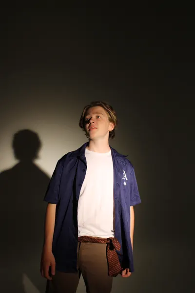 J.J. Barton looking upward in an open blue shirt over a white tee, his shadow cast on the wall beside him.