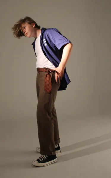 J.J. Barton leaning forward in an open blue short-sleeved shirt over a white tee, brown trousers and black high-tops, against a grey studio wall.