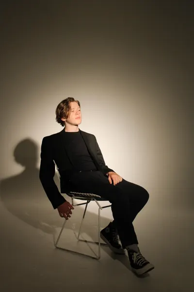 Wide shot of J.J. Barton on a folding chair, legs crossed, gazing upward in a black suit.