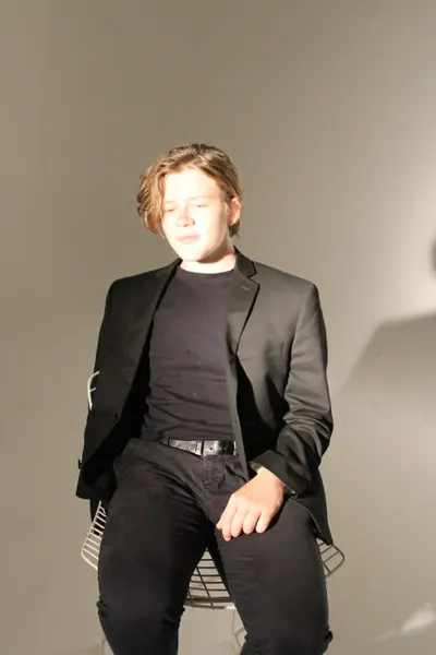 J.J. Barton sitting on a wire chair in a black tee and blazer, eyes closed, hands on his knees.