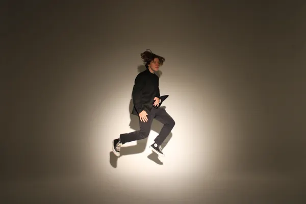 J.J. Barton leaping with one knee bent, in a dark suit, lit by a single spot on a plain wall.