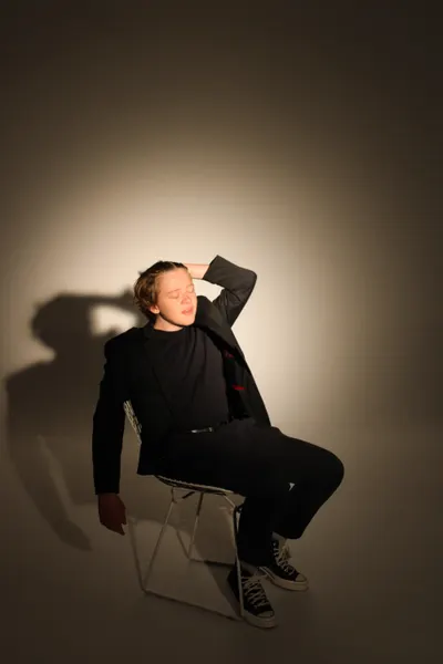 J.J. Barton reclining on a folding chair with one hand behind his head, legs crossed, in a black suit.
