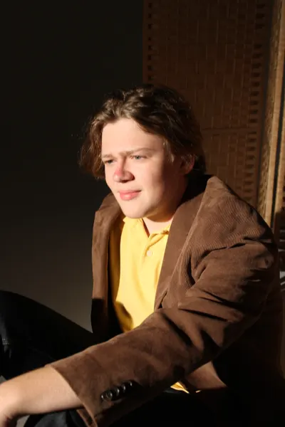 J.J. Barton sitting in a brown corduroy jacket and yellow polo shirt, looking away from the camera, lit from one side against a dark background.