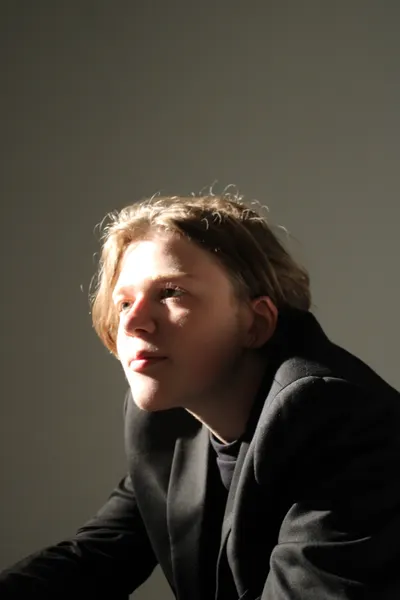 J.J. Barton leaning forward on his elbows in a dark jacket, half his face caught in a shaft of light.