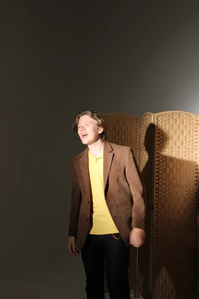 J.J. Barton standing and laughing with his head thrown back, in a brown jacket over a yellow polo shirt.