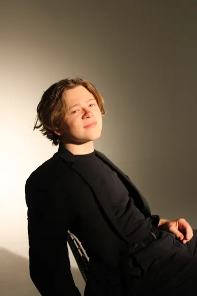 J.J. Barton seated with his head tilted, smiling at the camera, in a black suit and roll-neck.