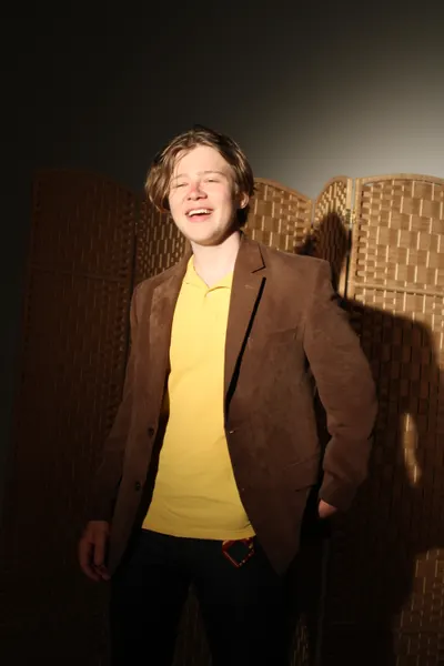 J.J. Barton laughing with his eyes closed and one hand in his pocket, in a brown jacket and yellow polo shirt.