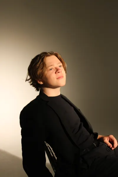 J.J. Barton leaning back in a chair looking upward, half-smiling, in a black suit and roll-neck.