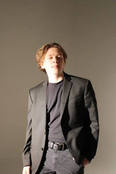J.J. Barton standing with one hand in his pocket, in a grey blazer over a dark tee and jeans.