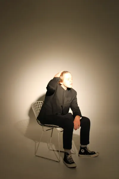 Wide shot of J.J. Barton on a wire chair, one hand behind his head and eyes closed, in a dark suit.