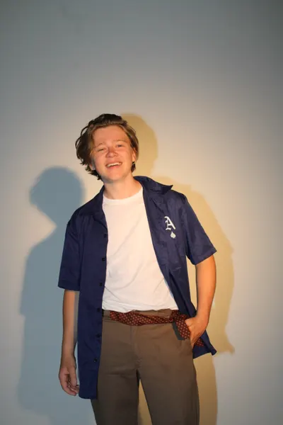 J.J. Barton laughing with one hand in his pocket, in an open blue shirt over a white tee and brown trousers.