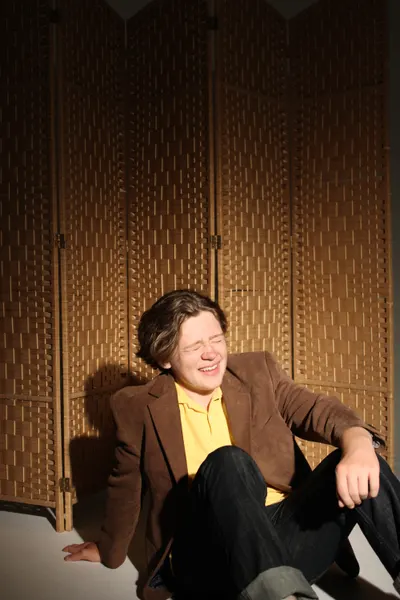 J.J. Barton sitting on the floor against a woven screen, head back, laughing with his eyes shut.