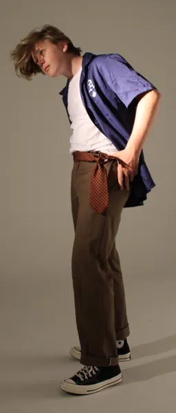 J.J. Barton bent forward at the waist, hair falling across his face, in an open blue shirt, brown trousers and black canvas high-tops.