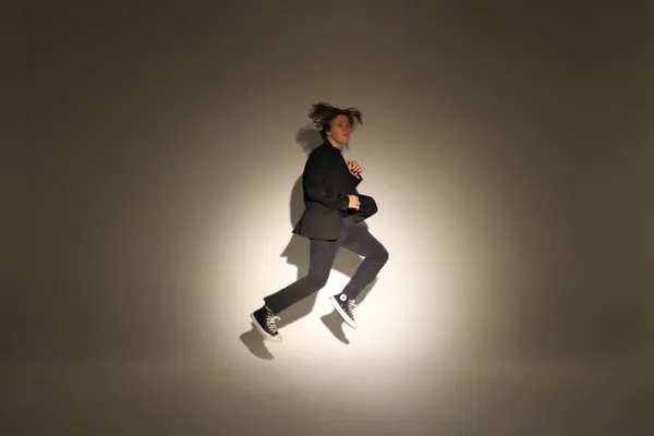 J.J. Barton in mid-air with his hair flying, wearing a dark suit, against a softly lit studio wall.
